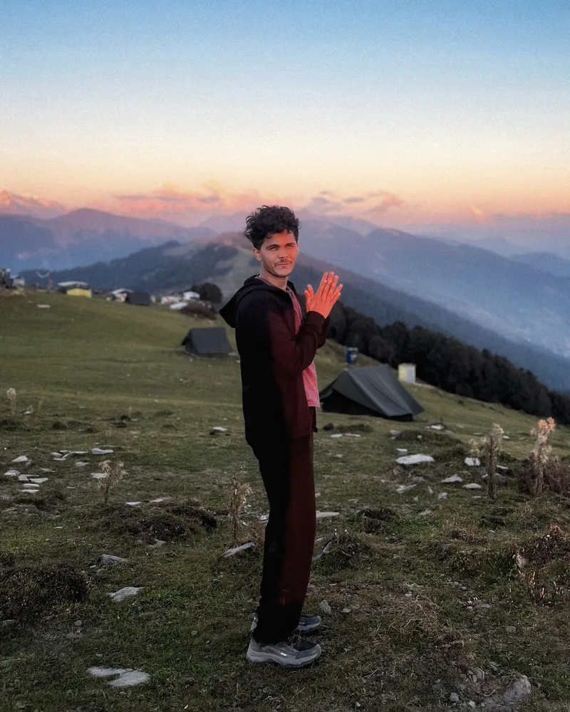 Ansith on a hillside at sunset, hands pressed together, with tents and mountains behind
