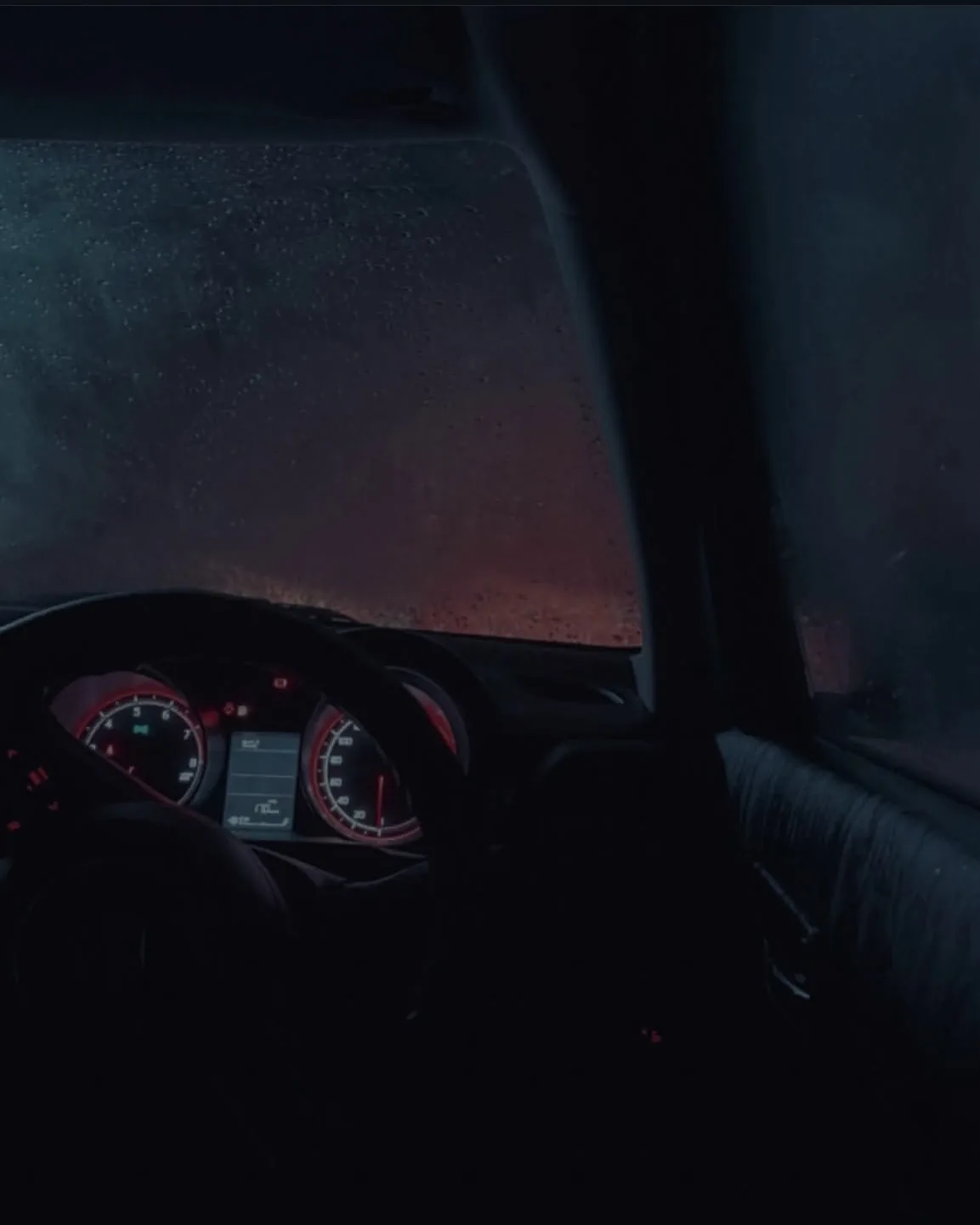 A dark car interior with glowing red dials and a rain-covered windshield