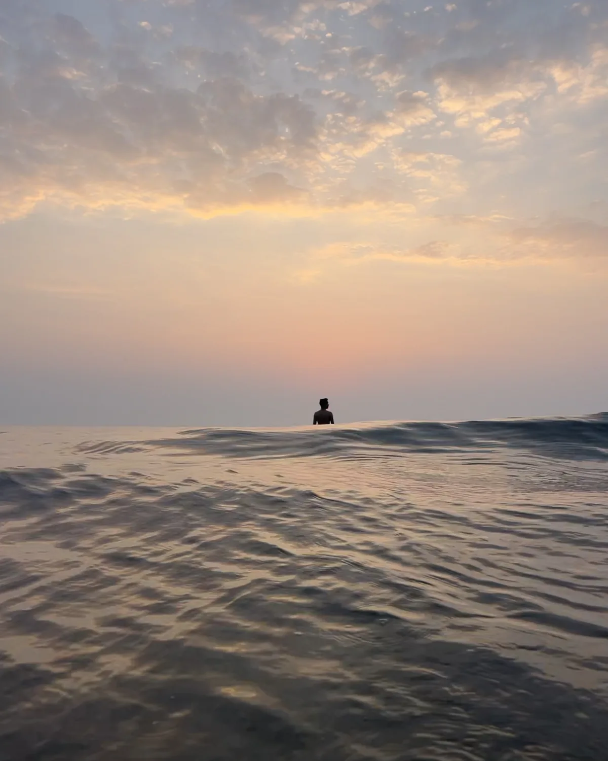 A silhouette in calm water facing a peach and lilac sunset