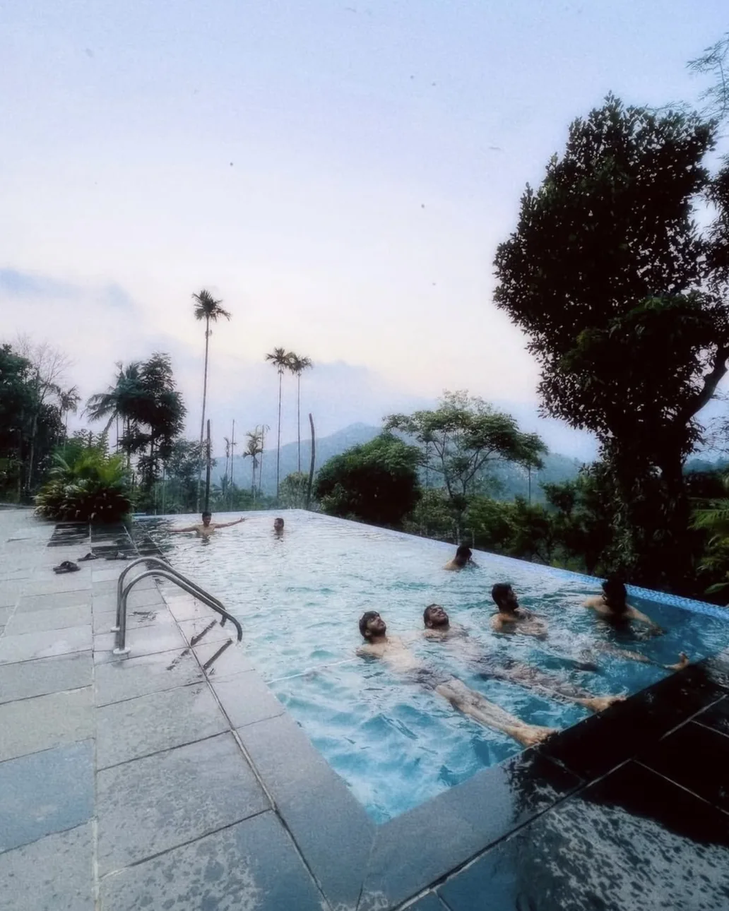 Friends floating in an infinity pool above a forested valley at dusk